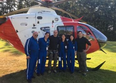 6 people standing in front of a red and white helicopter sitting on a field. 