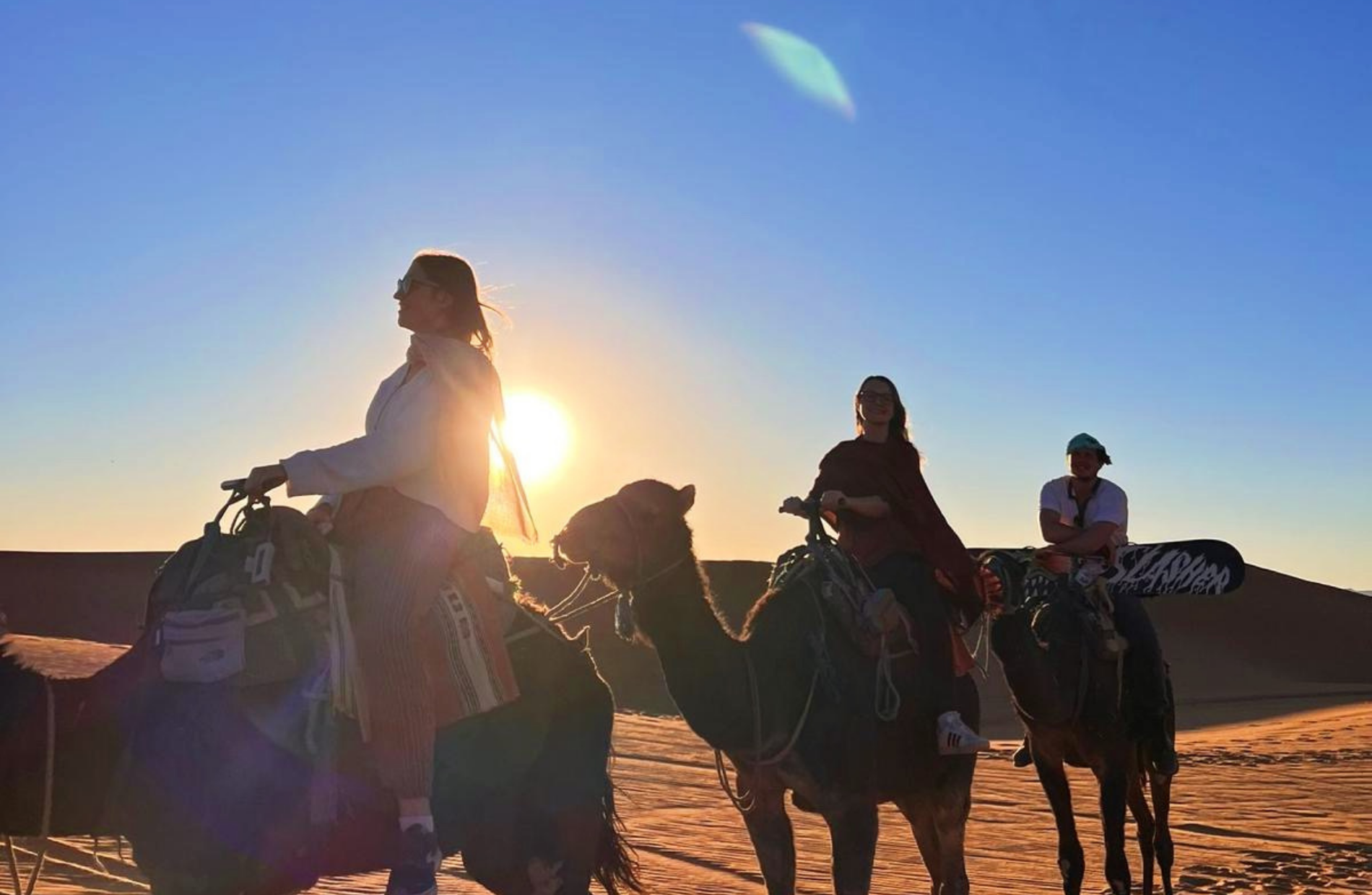 three people atop three camels with sand dunes in the back on a blue sky sunny day