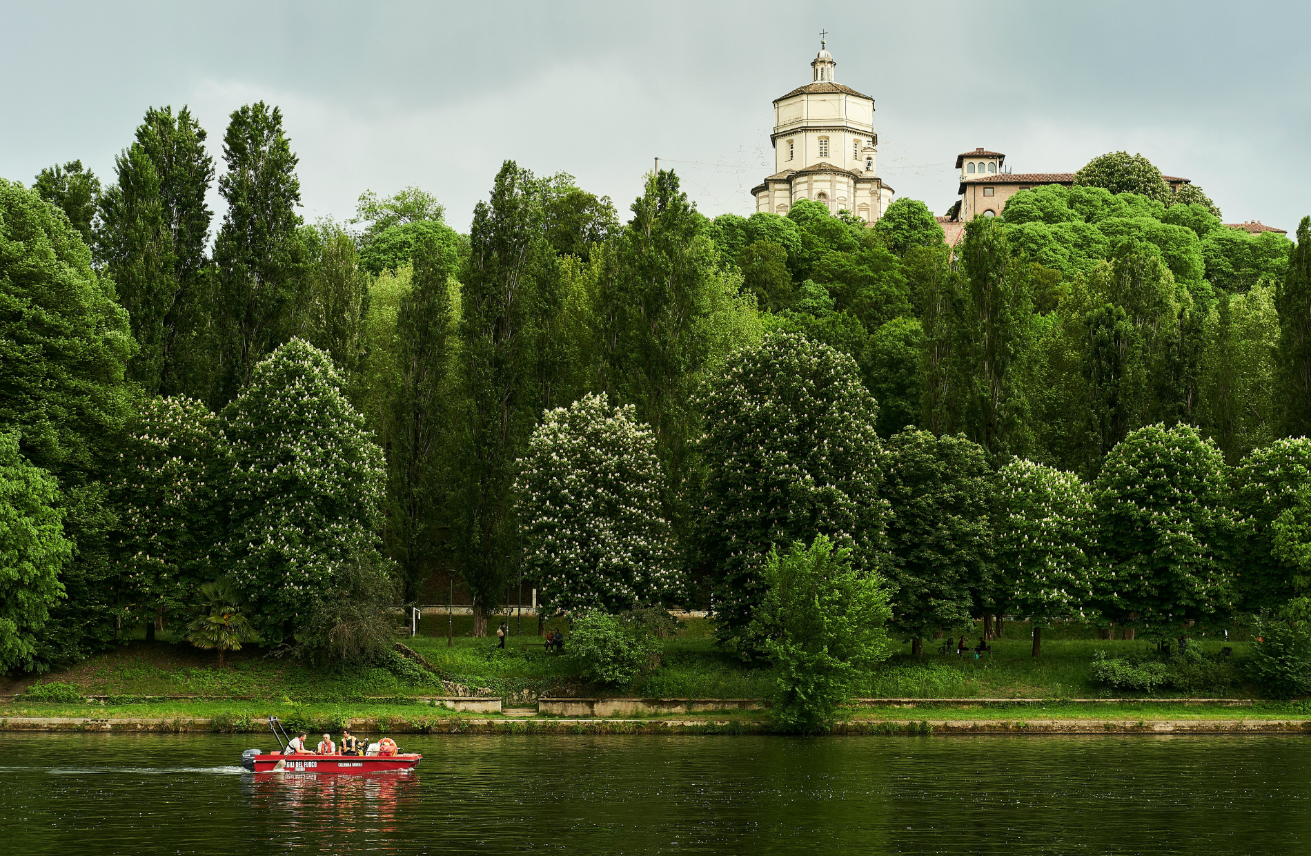 view of people riding in a boat on water with lush green trees in the background and a medieval tower peeking from behind the trees