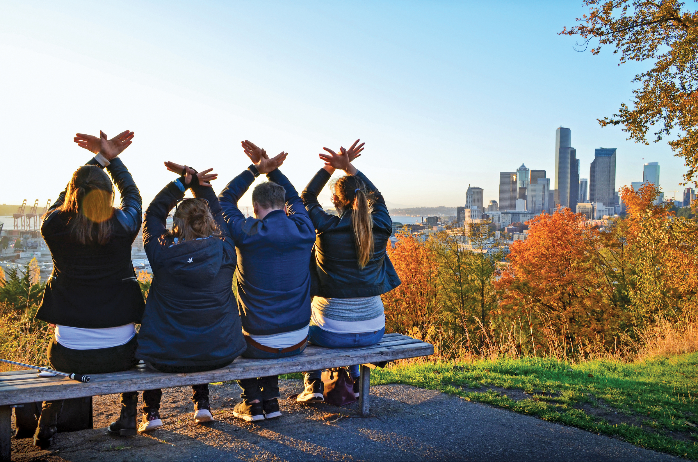 four students sitting on a bench overlooking the city skyline with hawk hands overhead