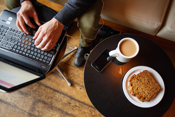 Hand typing on a laptop next to their coffee, pastry, and phone